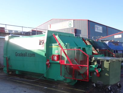 A Gradeall GPC P24 wet waste compactor, shown lifting a 1100 litre wheelie bin, with a red safety cage for secure operation
