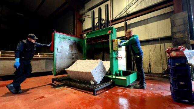 Two workers operating an aluminium can baler as it ejects a compressed bale of cans.