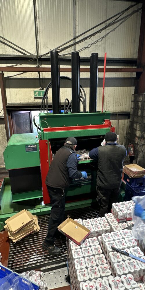 Workers operating a Gradeall Can Baler amidst an array of aluminium cans ready for recycling.