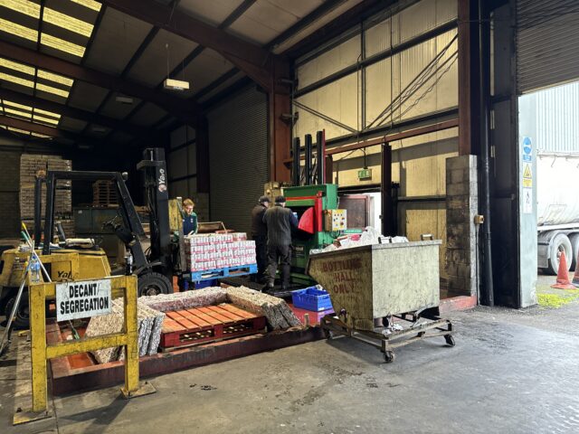 Workers at a beverage can recycling facility operate a Gradeall can baler beside a forklift and segregated waste.