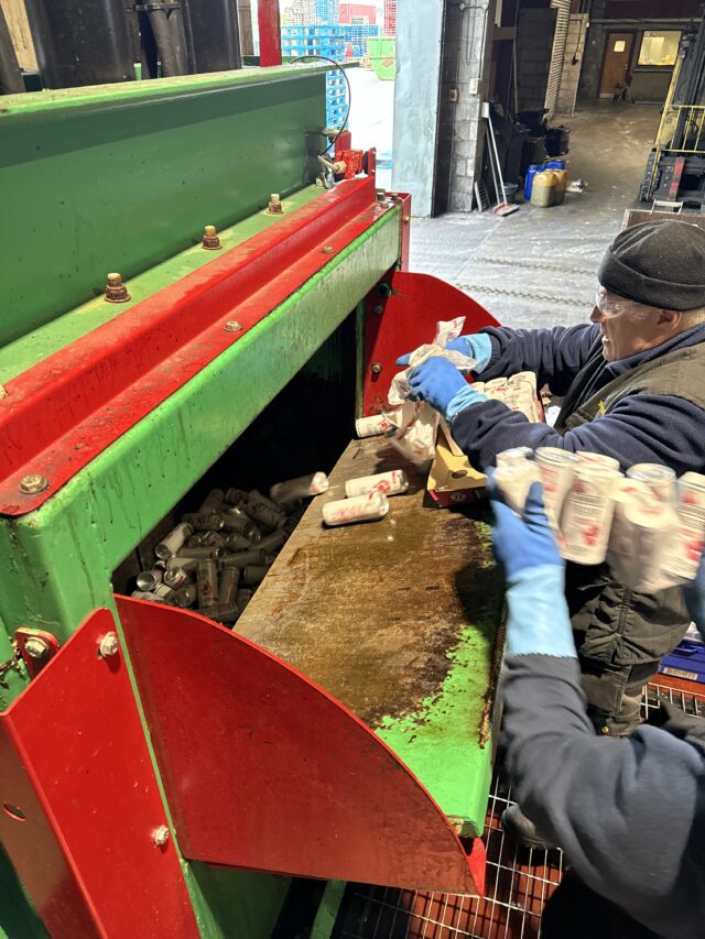 A worker feeds beverage cans into the hopper of a Gradeall can baler at a recycling facility.