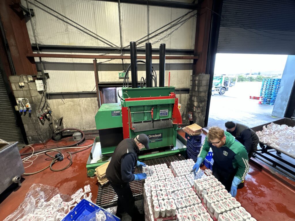 Workers sorting soda cans for recycling with a Gradeall can baler machine in the background