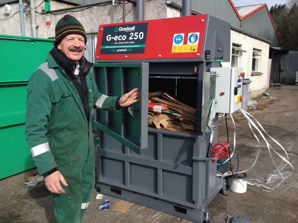 Worker smiling next to a Gradeall G-Eco 250 mid-size vertical baler filled with cardboard